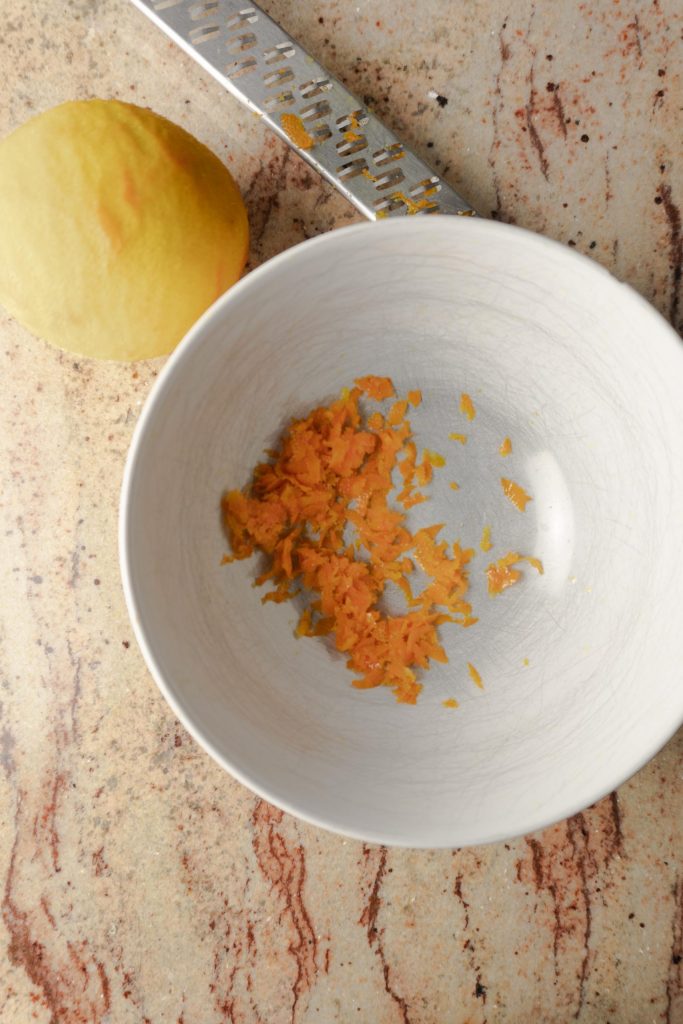 Orange zest in a white bowl with a grater and an orange in background