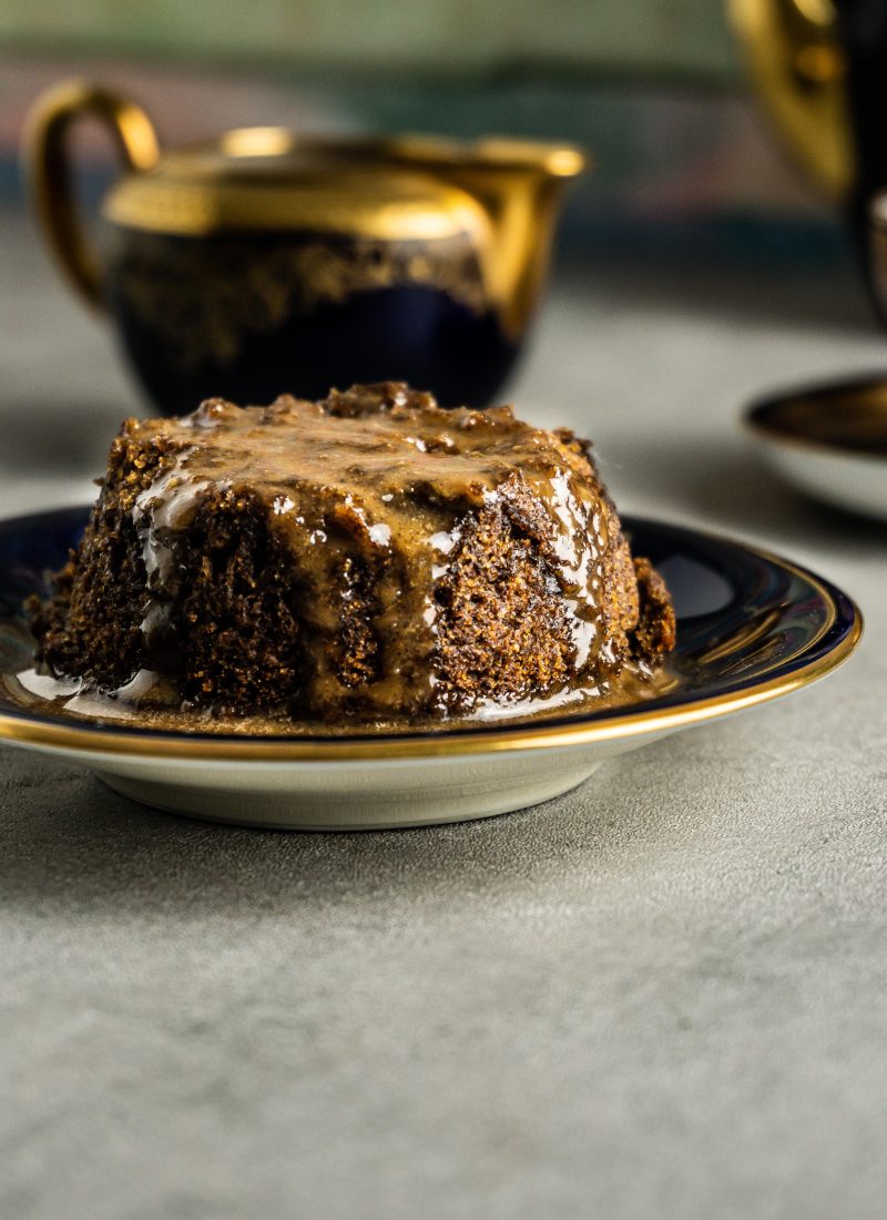 Vegan Hot Sticky Toffee Pudding on a small plate with sticky toffee pudding sauce and a saucer in the background