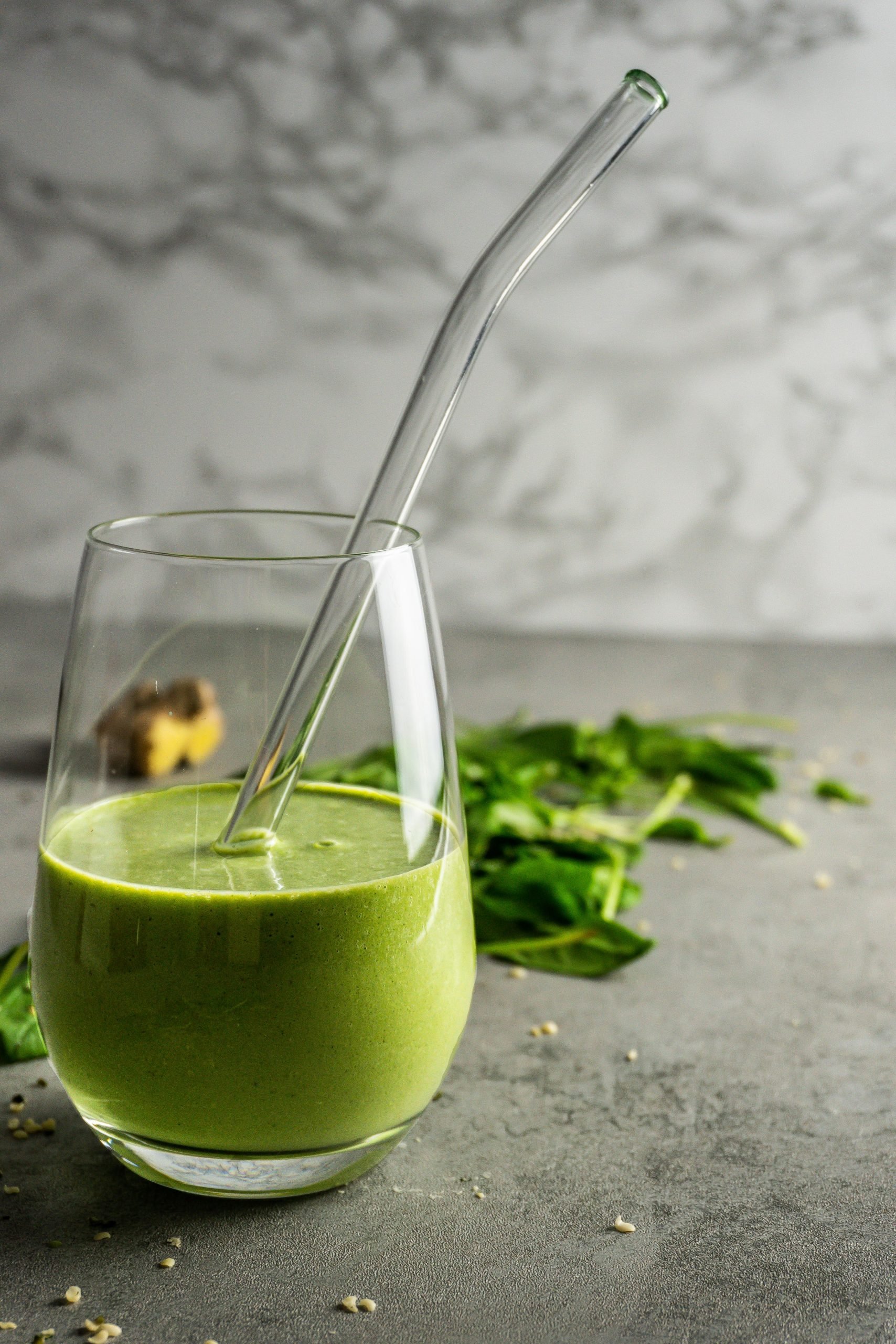 Green Monster Smoothie in a glass with a glass straw. Photographed on a grey background with scattered spinach and hemps seeds in the background