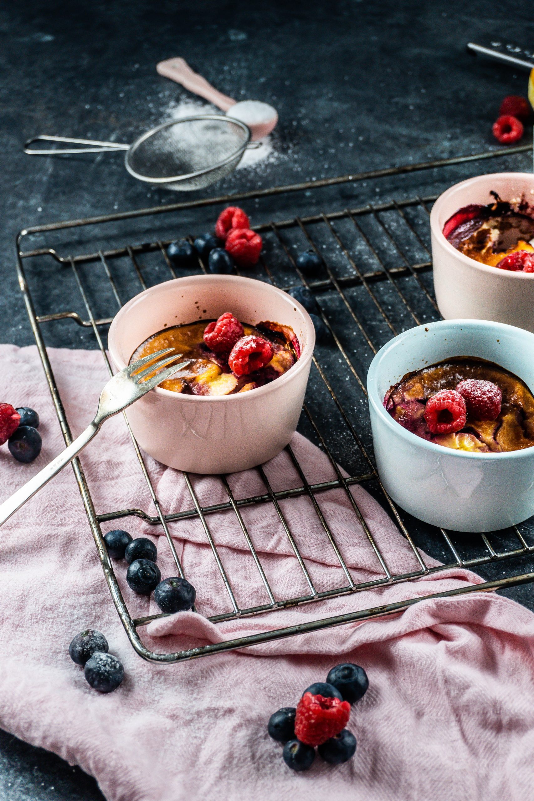 Five Minute Mini Cheesecakes on a baking tray surrounded with raspberries and blueberries covered with some powdered sugar and a fork tugging into one of the mugs