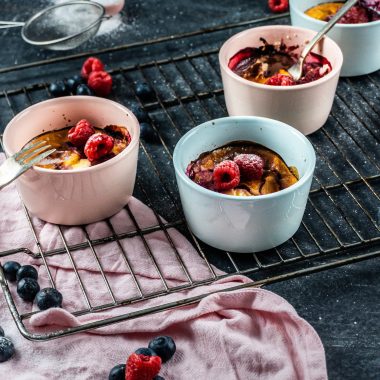 Five Minute Mini Cheesecakes on a baking tray surrounded with raspberries and blueberries covered with some powdered sugar and forks tugged into two mugs