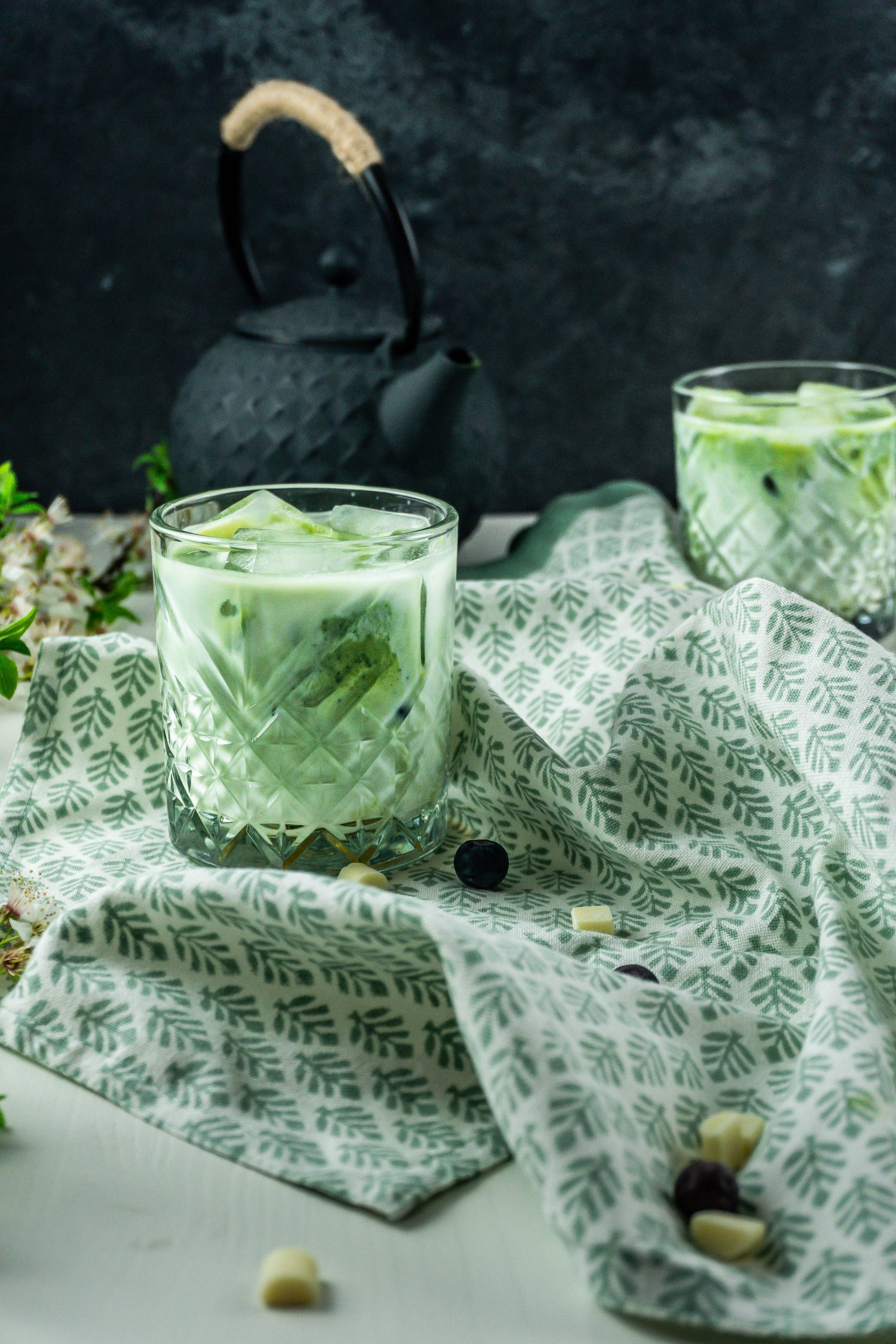 Iced Matcha Lattes in a glass, photographed on a kitchen towel with a tea pot in the background