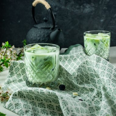 Iced Matcha Lattes in a glass, photographed on a kitchen towel with a tea pot in the background