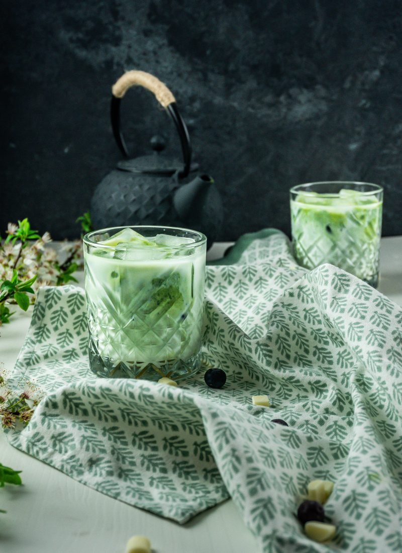 Iced Matcha Lattes in a glass, photographed on a kitchen towel with a tea pot in the background