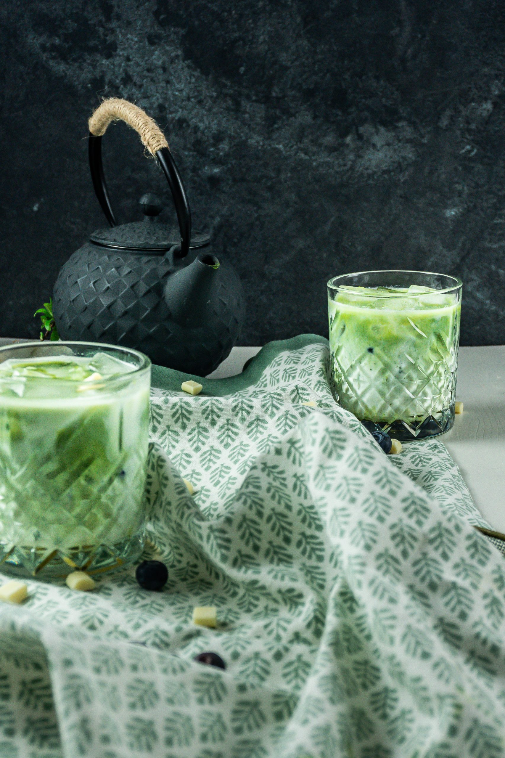 Iced Matcha Lattes in a glass, photographed on a kitchen towel with a tea pot in the background