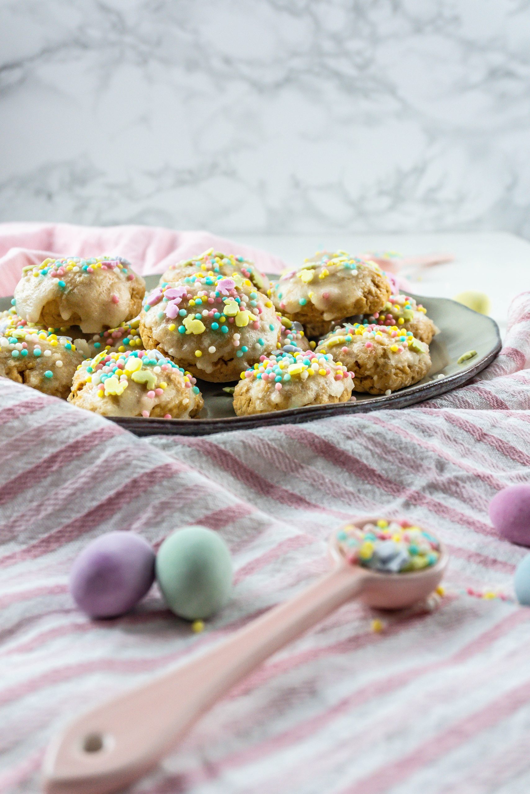 Italian Easter Cookies on a plate, surrounded by sprinkles, pastel Easter eggs and some dish towels; photographed from the side