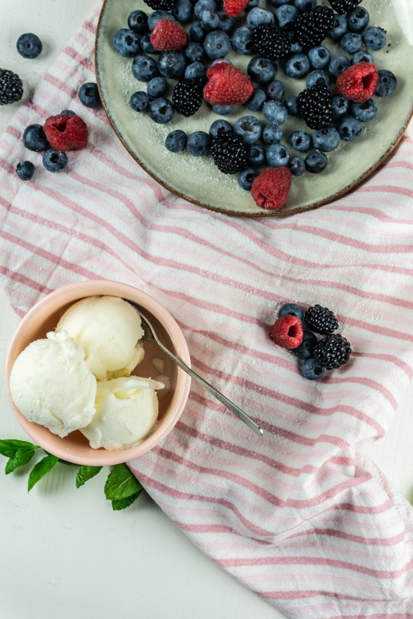Cool and creamy yogurt ice cream scoops with a spoon in it surrounded by mixed berries and some powdered sugar; photographed from above