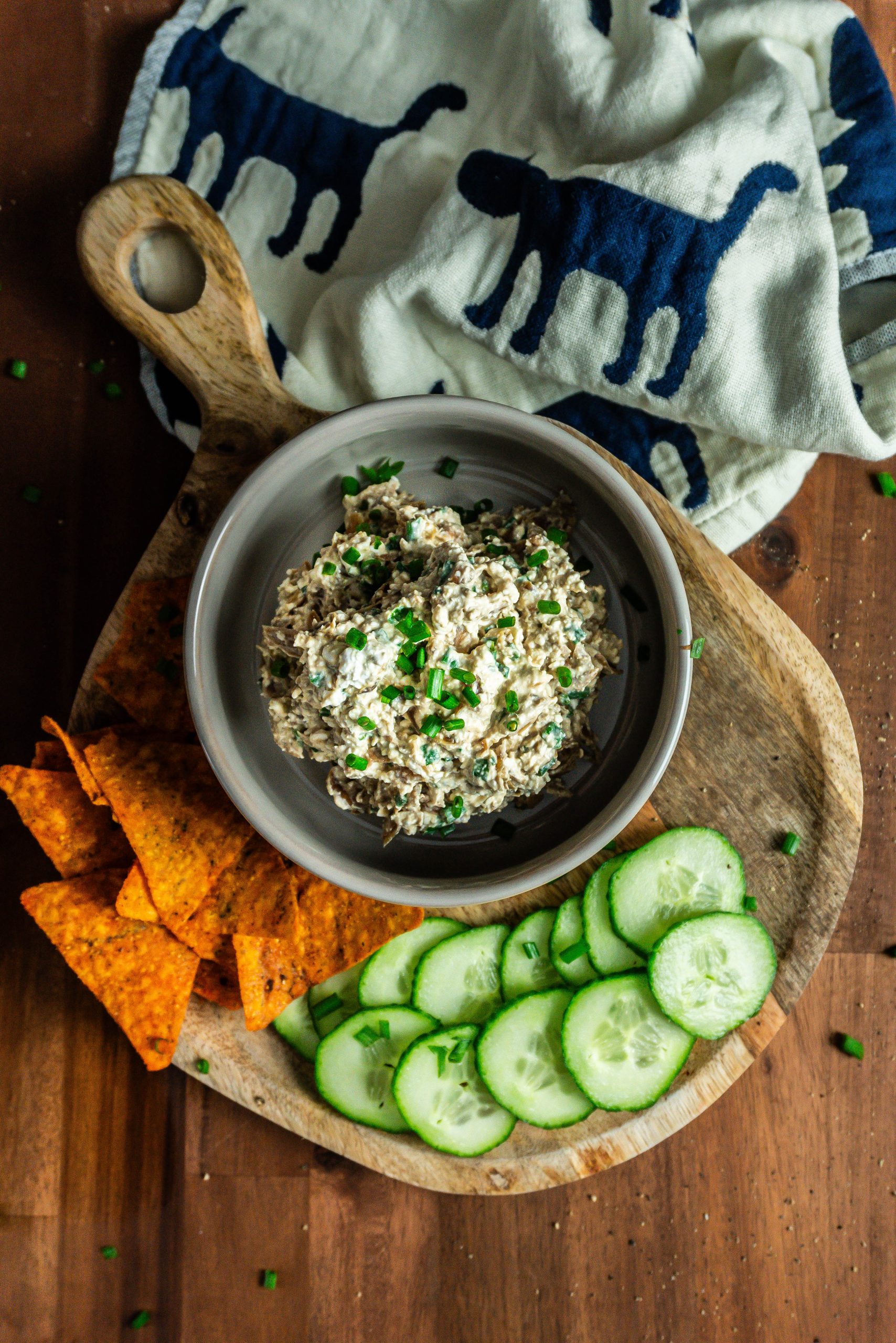Crowd-Pleasing Caramelized onion dip with cucumbers and nachos laying on the side on a small wooden board and chives strewn across