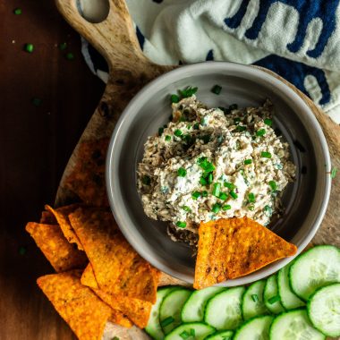 Crowd-Pleasing Caramelized onion dip with cucumbers and nachos laying on the side on a small wooden board and one nacho being stuck into the dip