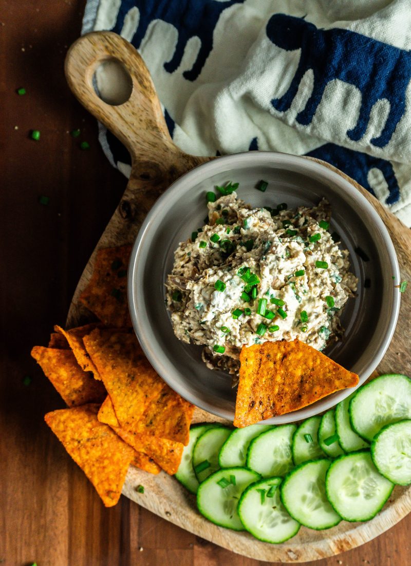 Crowd-Pleasing Caramelized onion dip with cucumbers and nachos laying on the side on a small wooden board and one nacho being stuck into the dip