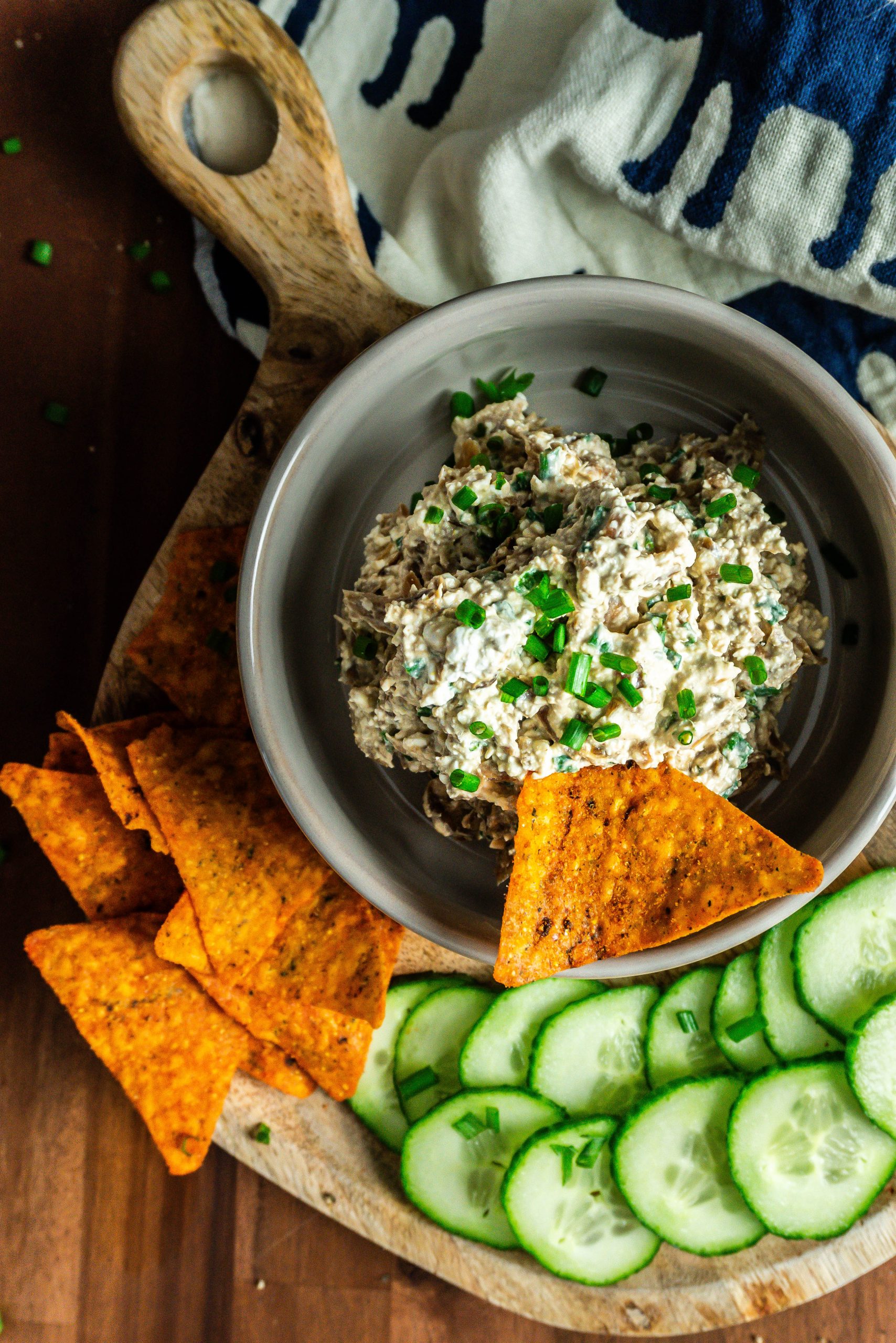 Crowd-Pleasing Caramelized onion dip with cucumbers and nachos laying on the side on a small wooden board and one nacho being stuck into the dip