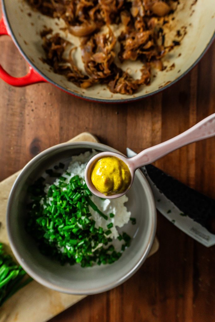 Mustard in a small spoon being in focus and being added to the crowd-pleasing caramelized onions dip