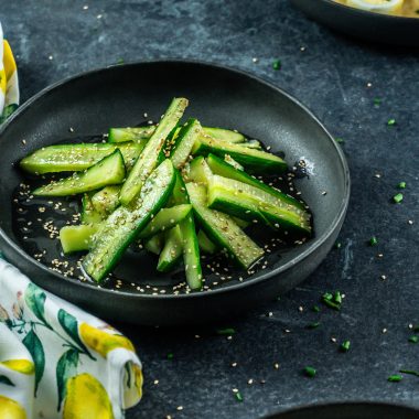 Japanese-Inspired Cucumber Salad on a black plate with other plates and some scattered sesame seeds visible in the background