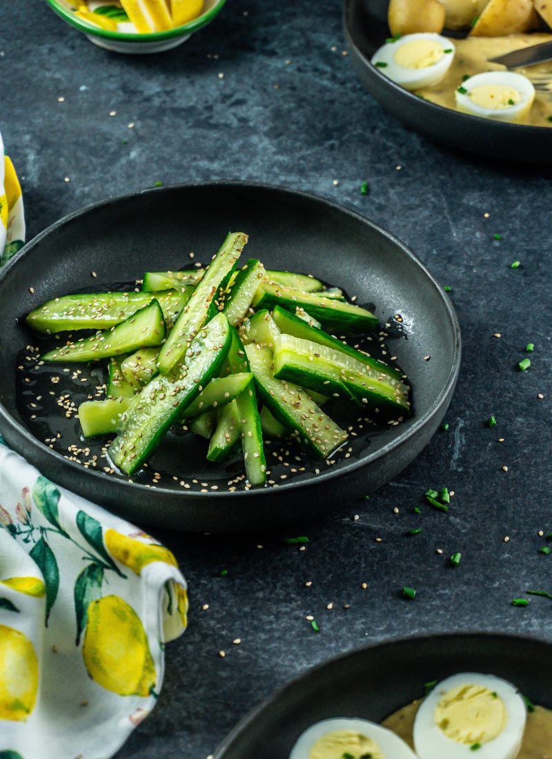 Japanese-Inspired Cucumber Salad on a black plate with other plates and some scattered sesame seeds visible in the background