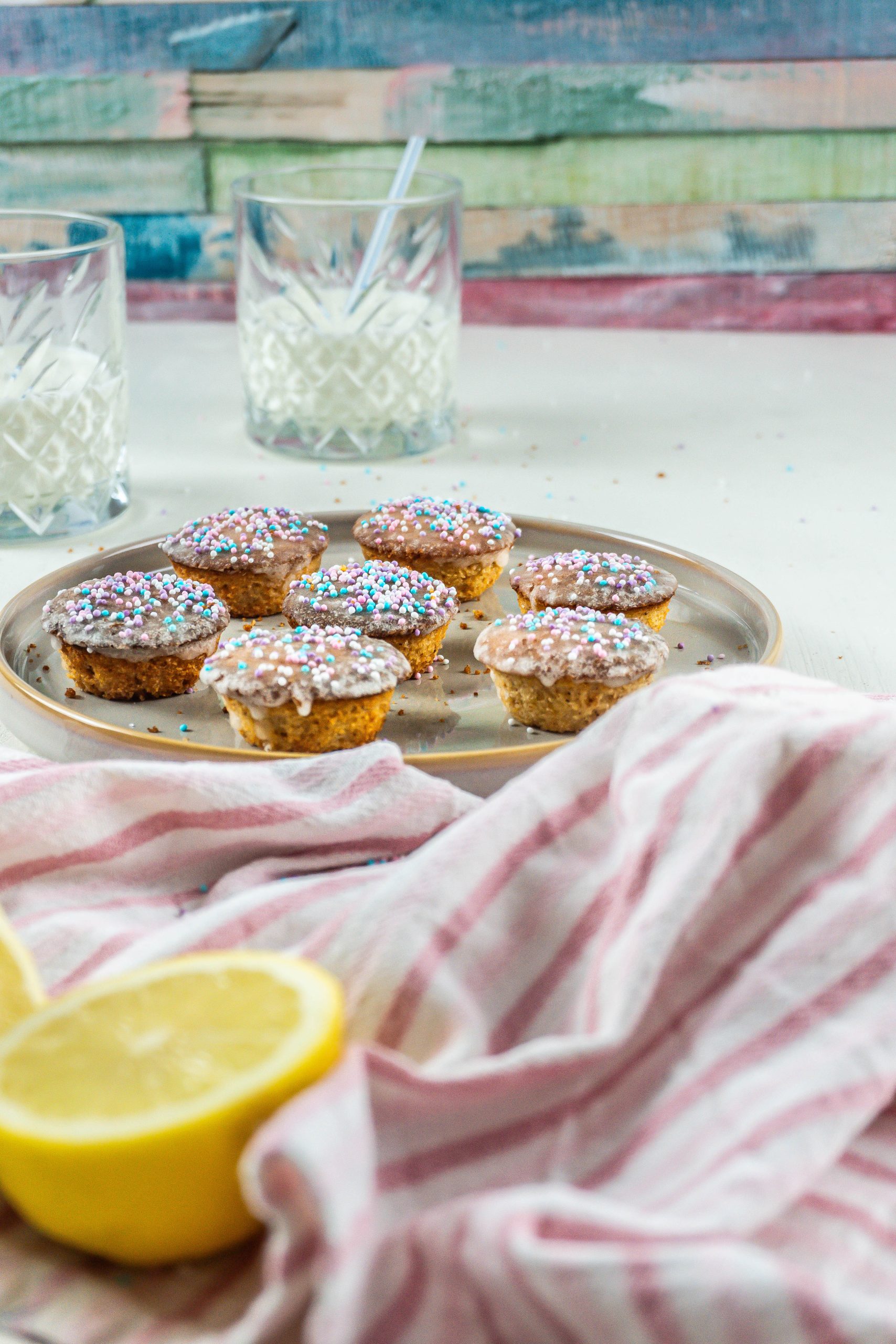 Lemon Mascarpone Mini Muffins on a small plate with two glasses of milk in the background and some sprinkles surrounding the plate and a lemon visible in the front of the frame