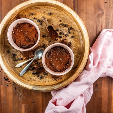 Gluten-free warm, fudgy mug brownies in in an oven-proof ramekin surrounded with chopped chocolate and cocoa powder
