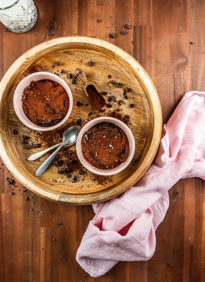Gluten-free warm, fudgy mug brownies in in an oven-proof ramekin surrounded with chopped chocolate and cocoa powder