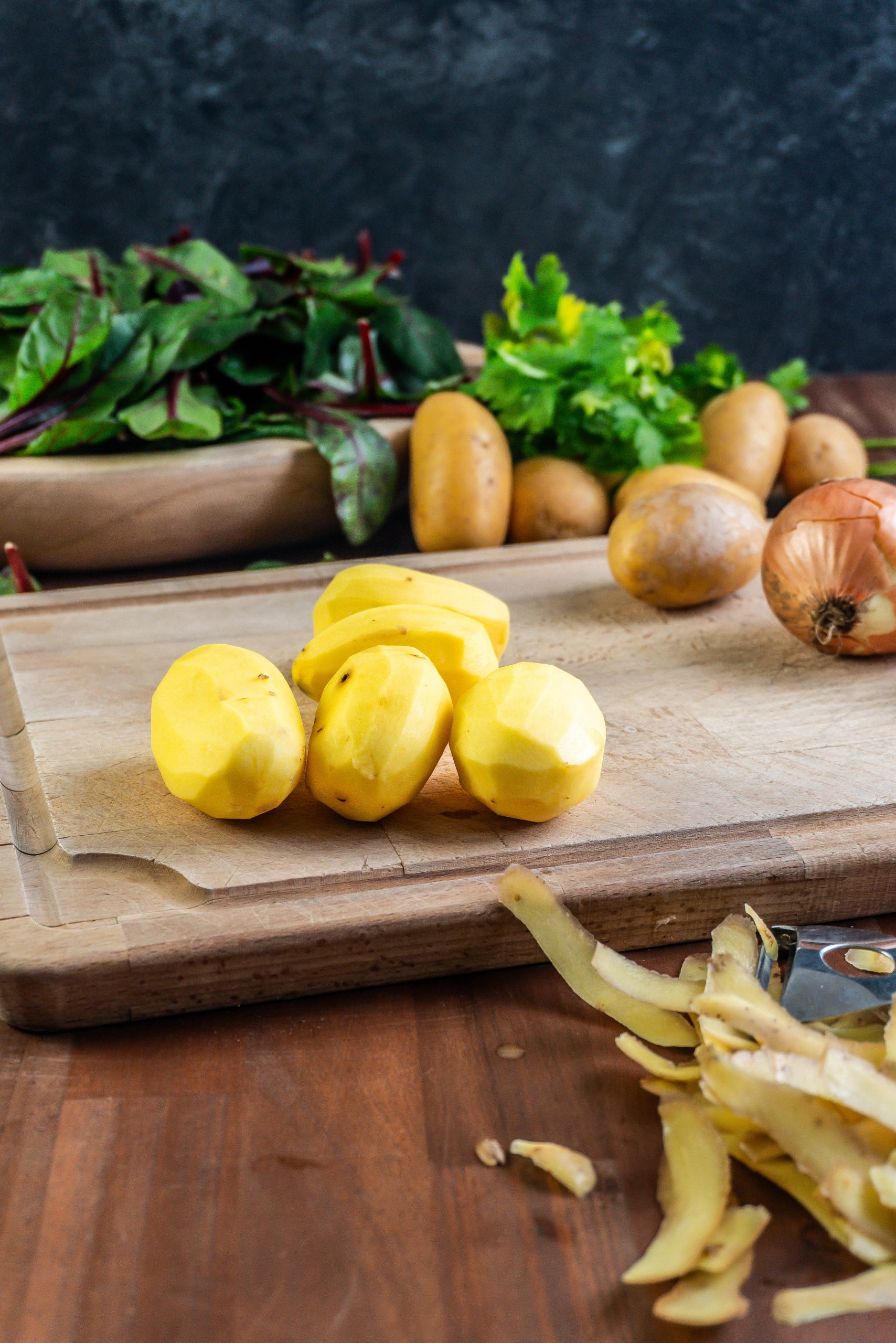 Peeled potatoes lying on a chopping board with young swiss chard visible in the background