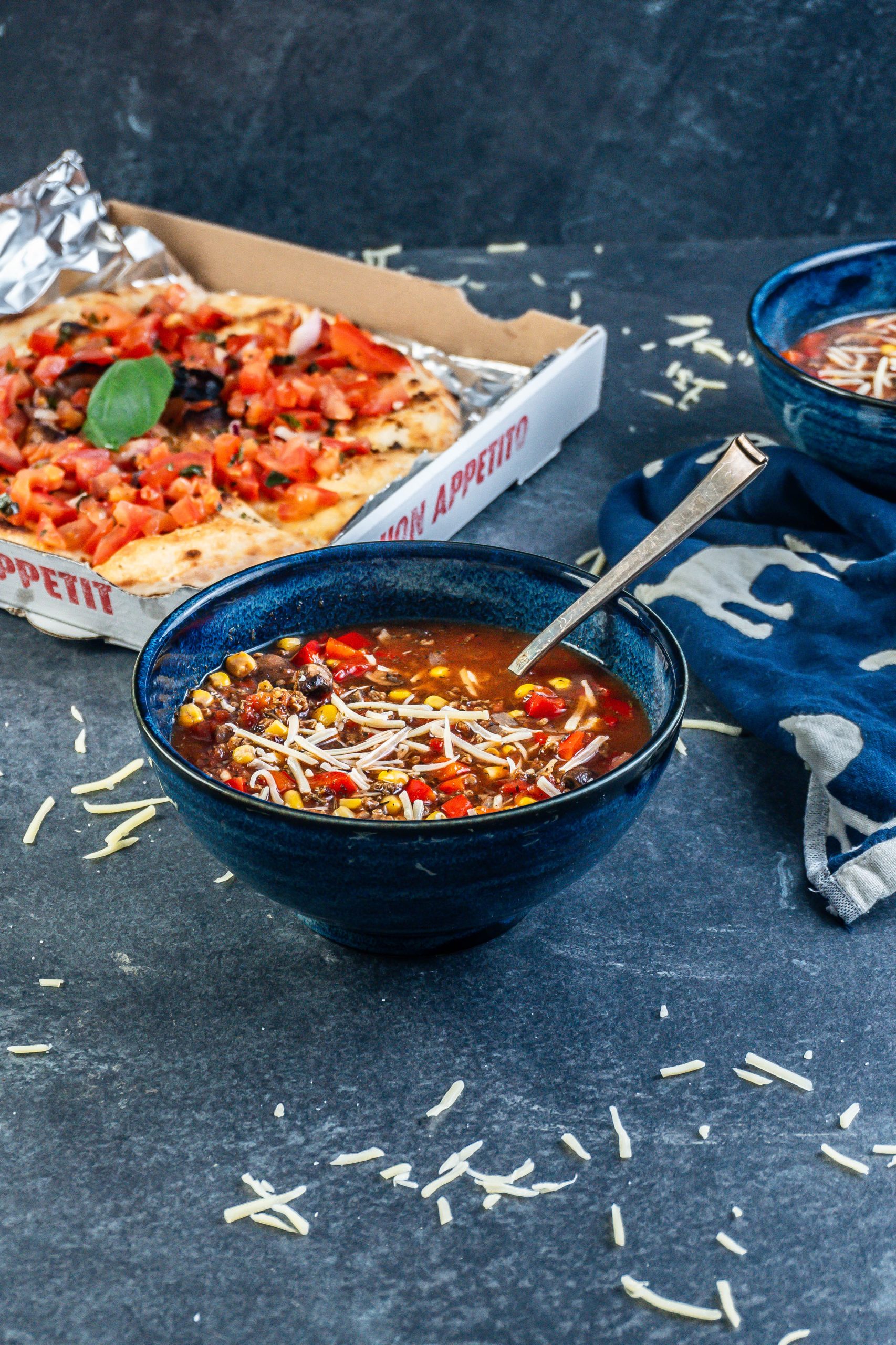 Hearty Vegan Pizza Soup Bowl decorated with some cheese and spoon stuck into the bowl; photographed from a side-angle