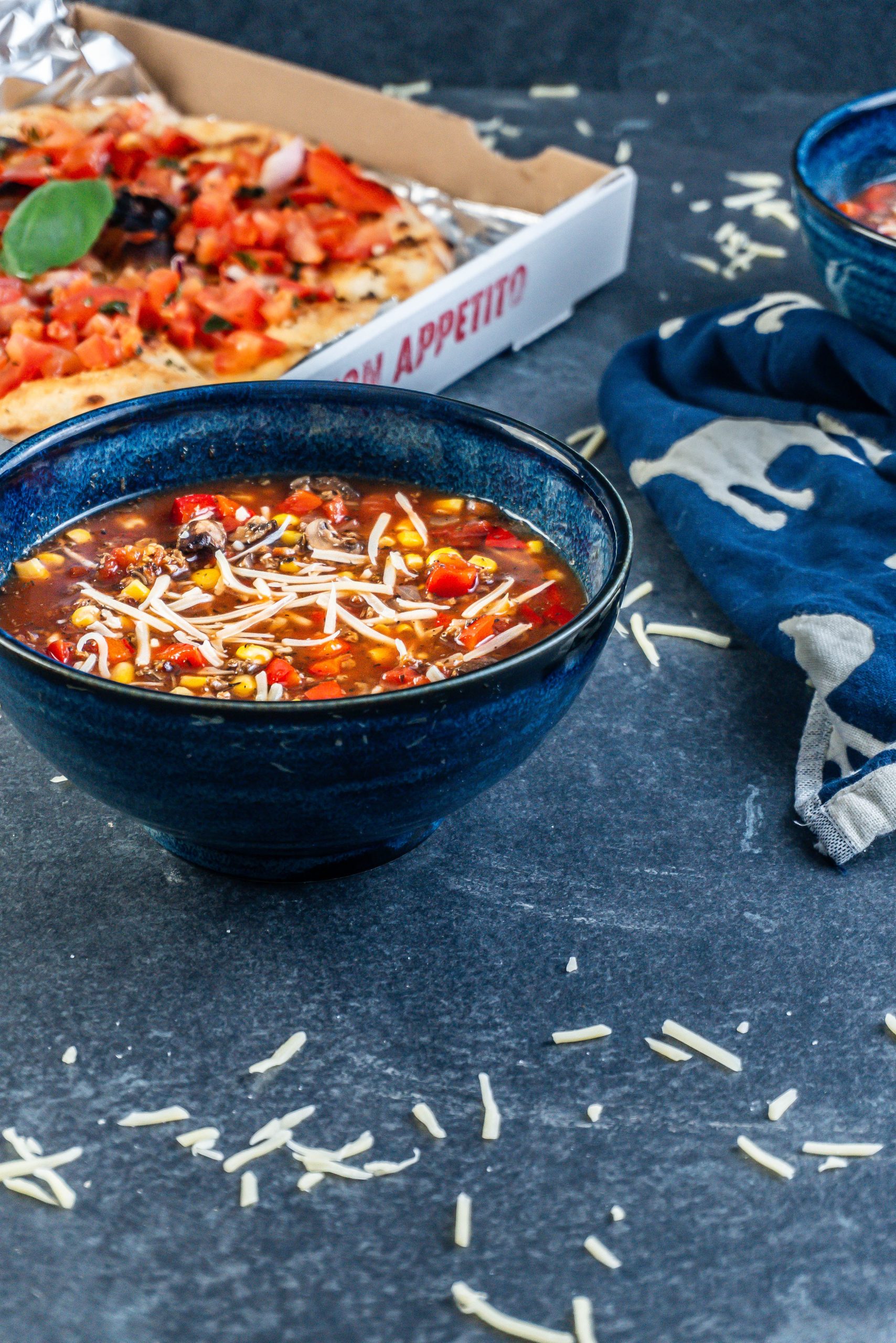 Hearty Vegan Pizza Soup Bowl decorated with some cheese and a pizza bread visible in the background; photographed from a side angle