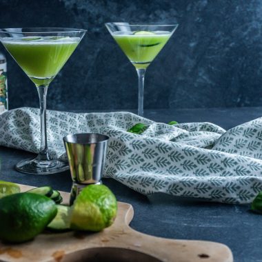Two non-alcoholic Garden Sour Cocktails in glasses with cucumber slices and juiced limes to the side; photographed from a side-angle