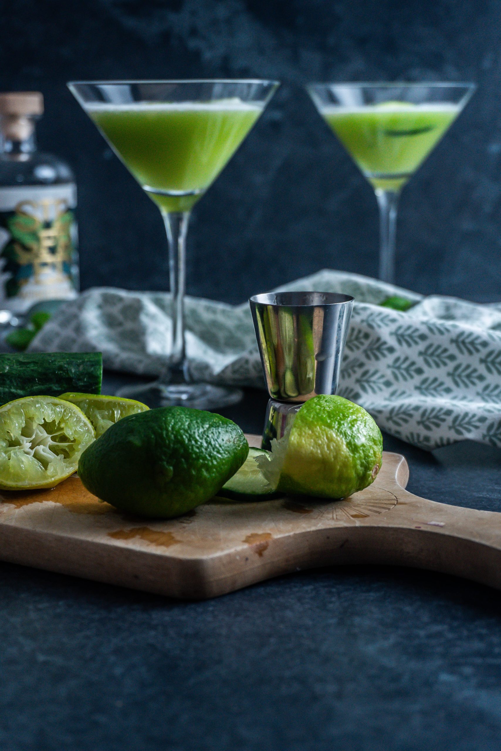 Juiced limes and cucumber slices in focus with two mocktails visible in the background