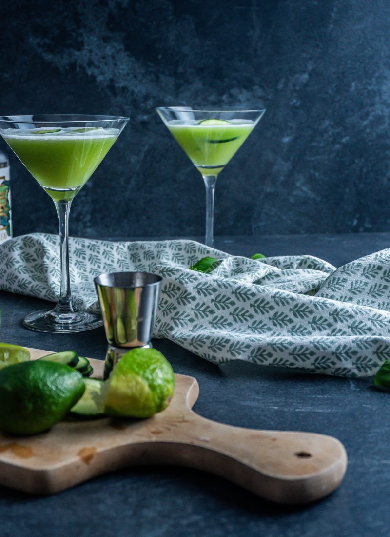Two non-alcoholic Garden Sour Cocktails in glasses with cucumber slices and juiced limes to the side; photographed from a side-angle