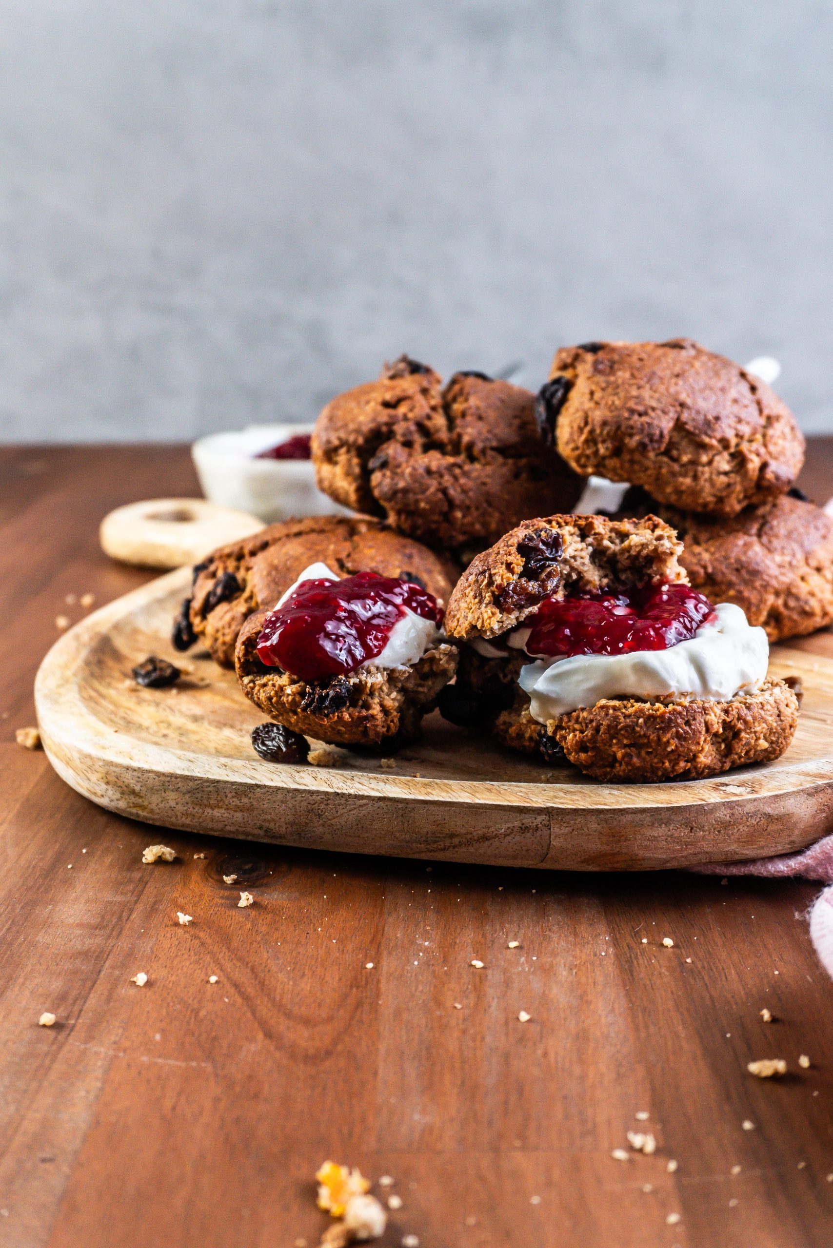 Cinnamon Orange Raisin Scones arranged on a platter with some jam and yogurt visible to the side and one scone being slathered in both; photographed from a side angle