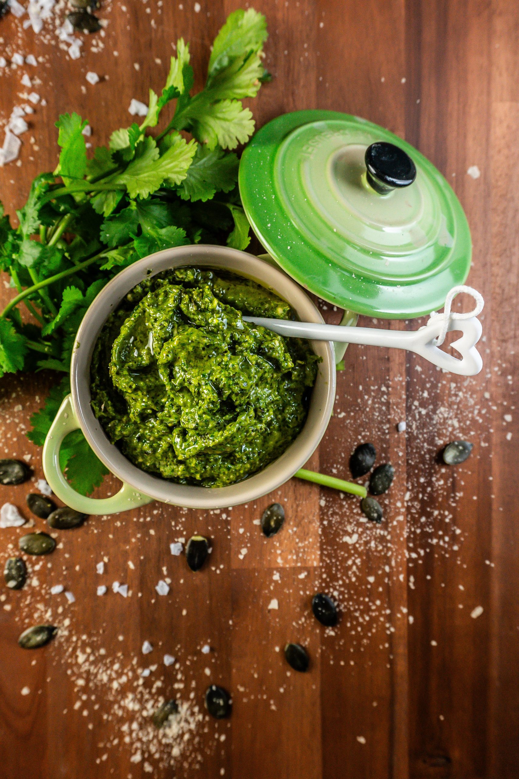 Five Minute Vegan Pepita Cilantro Pesto in a small ramekin dish with some cilantro, pepitas, and nutritional yeast scattered around and a spoon stuck into it