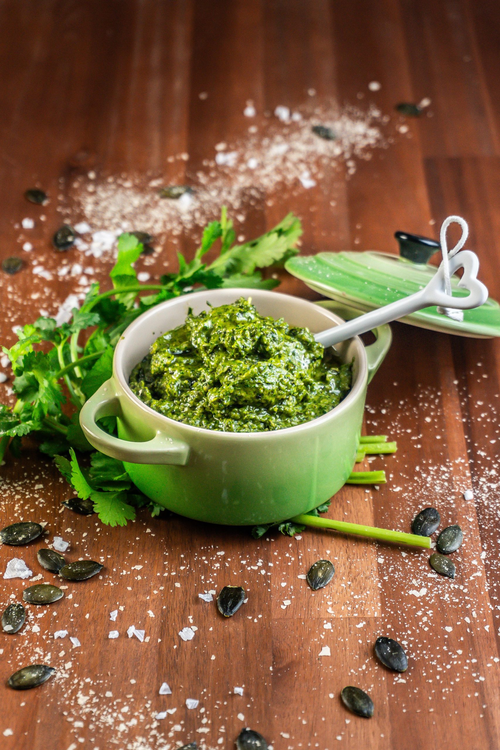 Five Minute Vegan Pepita Cilantro Pesto in a small ramekin dish with some cilantro, pepitas, and nutritional yeast scattered around and a spoon stuck into it