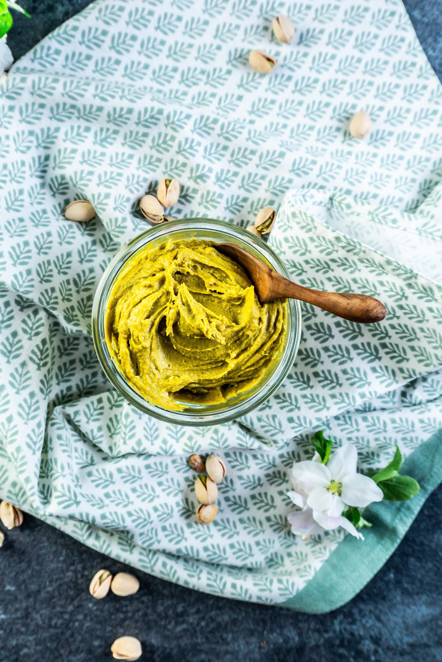 Pistachio nut butter in a glass jar with a wooden spoon stuck into it and some pistachios scattered around
