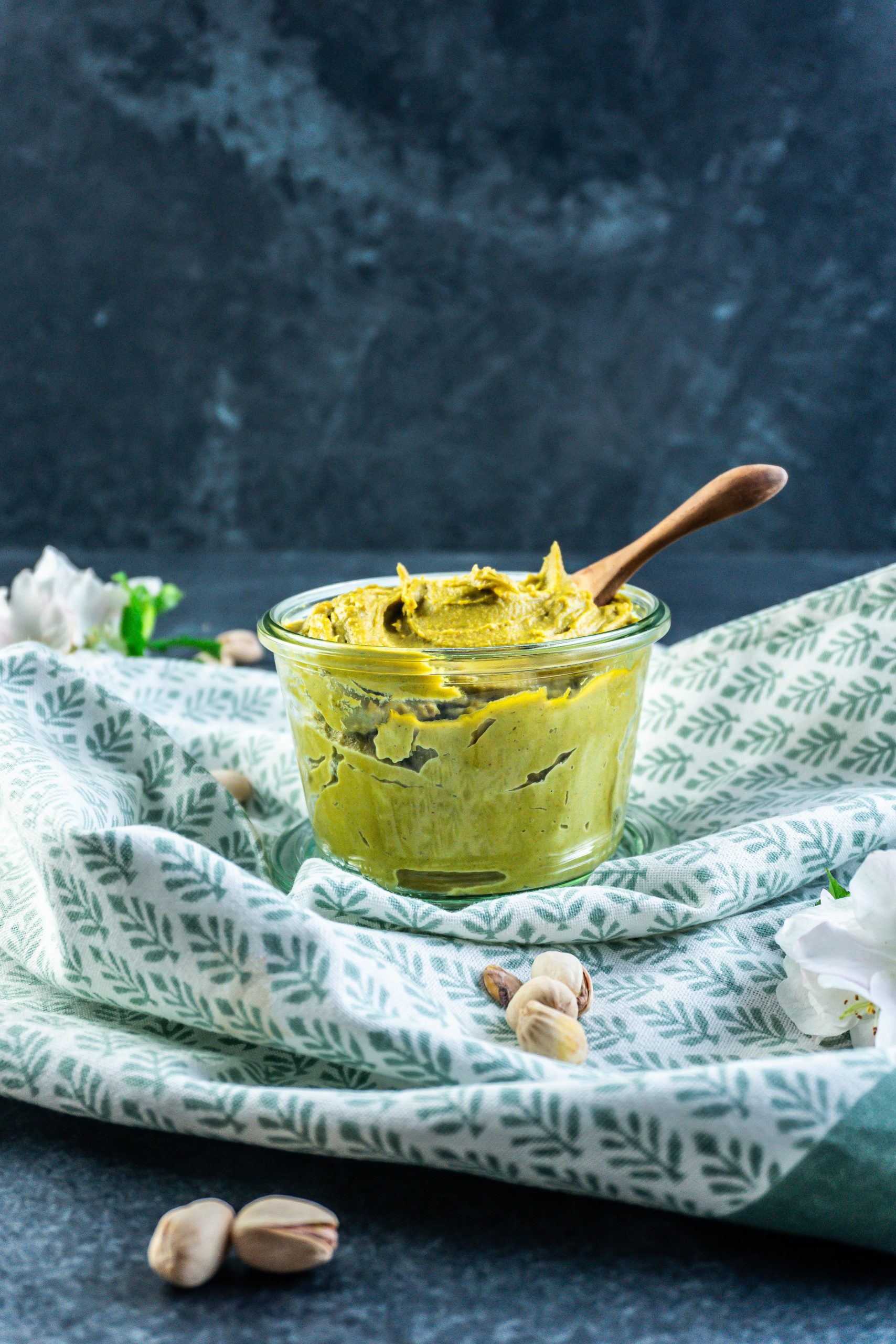 Pistachio nut butter in a glass jar with a wooden spoon stuck into it and some pistachios scattered around; photographed from a side angle