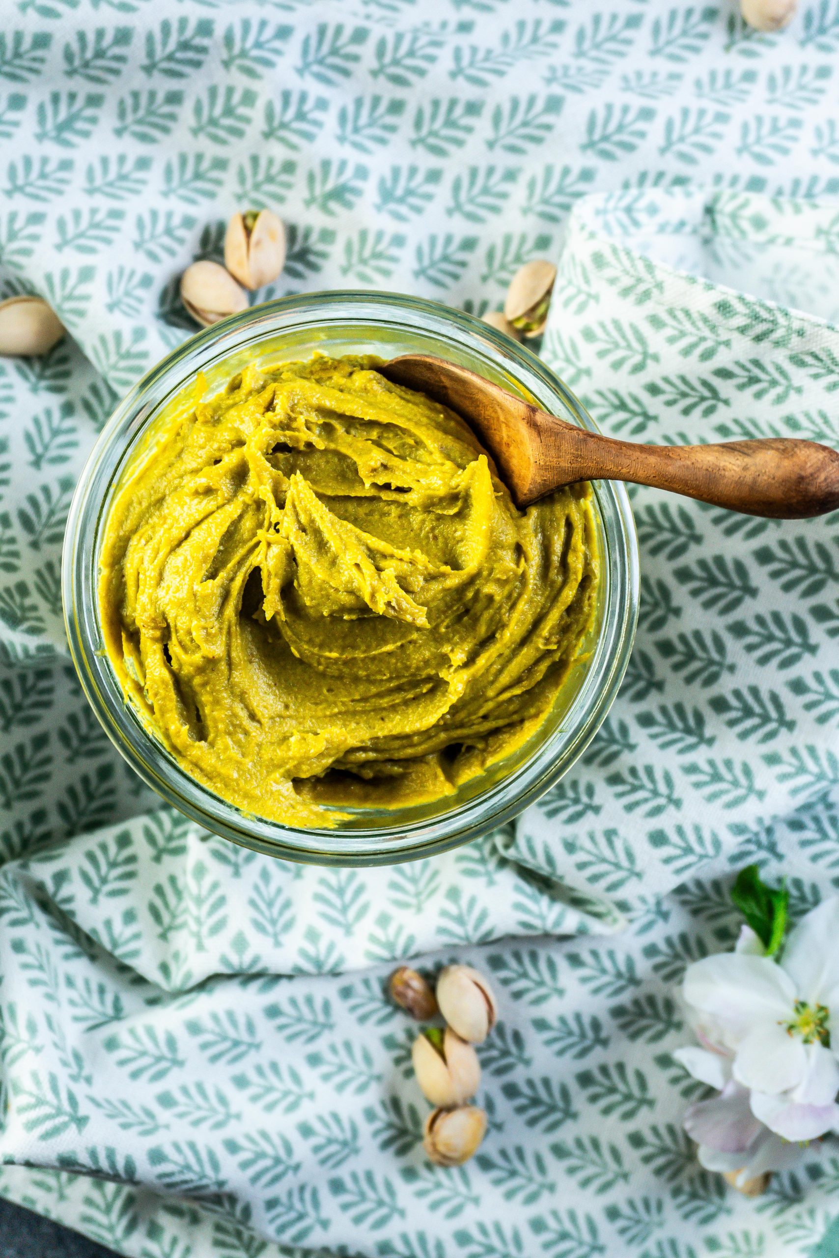 Pistachio nut butter in a glass jar with a wooden spoon stuck into it and some pistachios scattered around; photographed up close