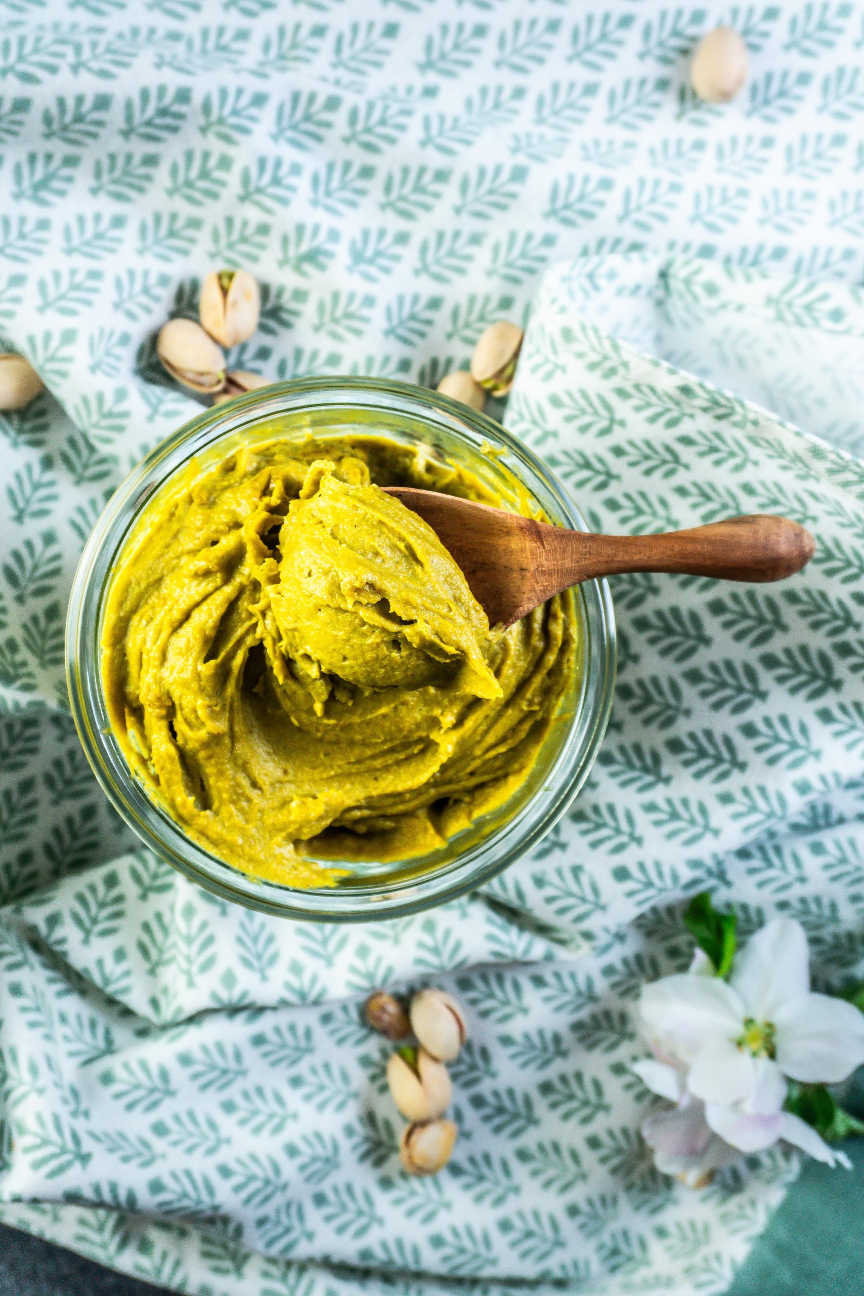 Pistachio nut butter in a glass jar with a wooden spoon scooping some nut butter on a spoon and some pistachios scattered around; photographed from above