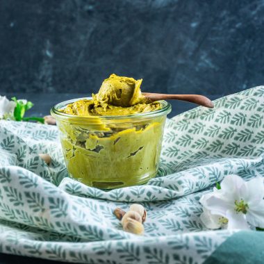 Pistachio nut butter in a glass jar with a wooden spoon scooping some nut butter on a spoon and some pistachios scattered around; photographed from a side angle