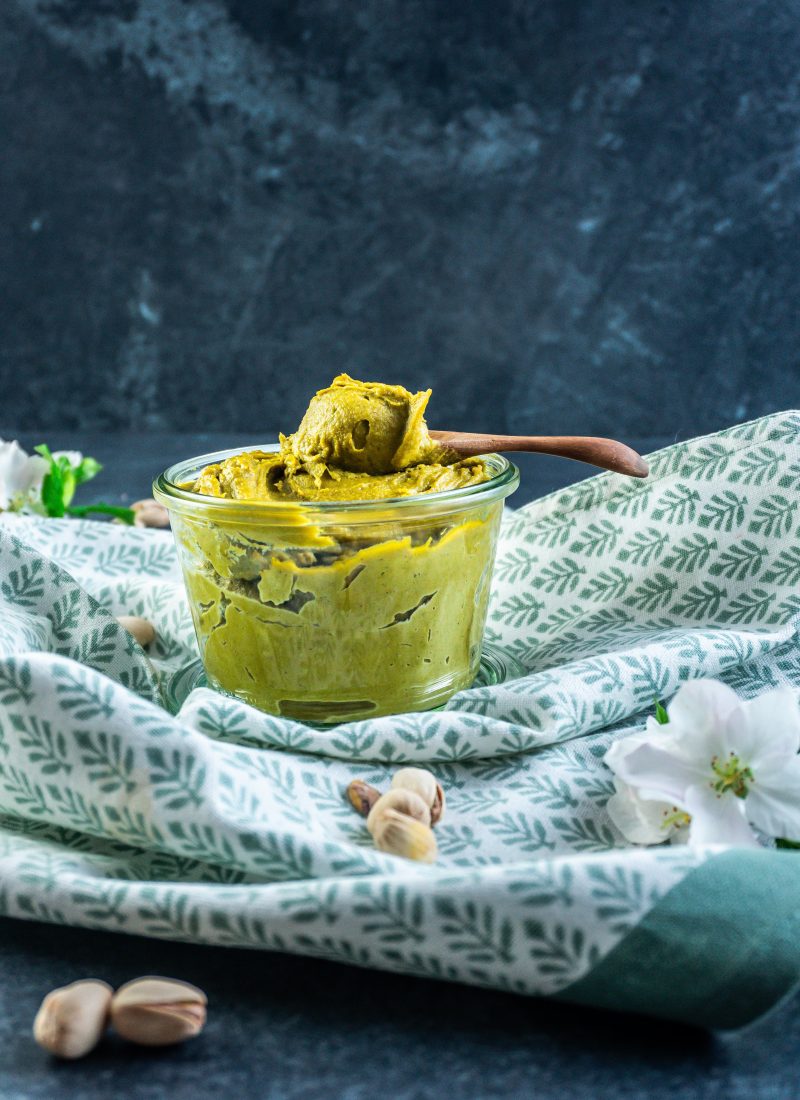 Pistachio nut butter in a glass jar with a wooden spoon scooping some nut butter on a spoon and some pistachios scattered around; photographed from a side angle