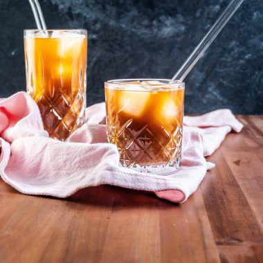 Coffee Ginger Elixir in a glass with a straw, another glass visible in the background on a kitchen towel