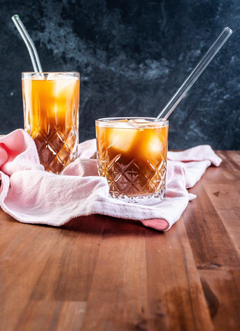 Coffee Ginger Elixir in a glass with a straw, another glass visible in the background on a kitchen towel