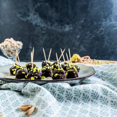 Gluten-free Dubai Style Pistachip Balls on a plate with some chopped pistachios strewn on top and some pistachios in a glass jar visible in the background