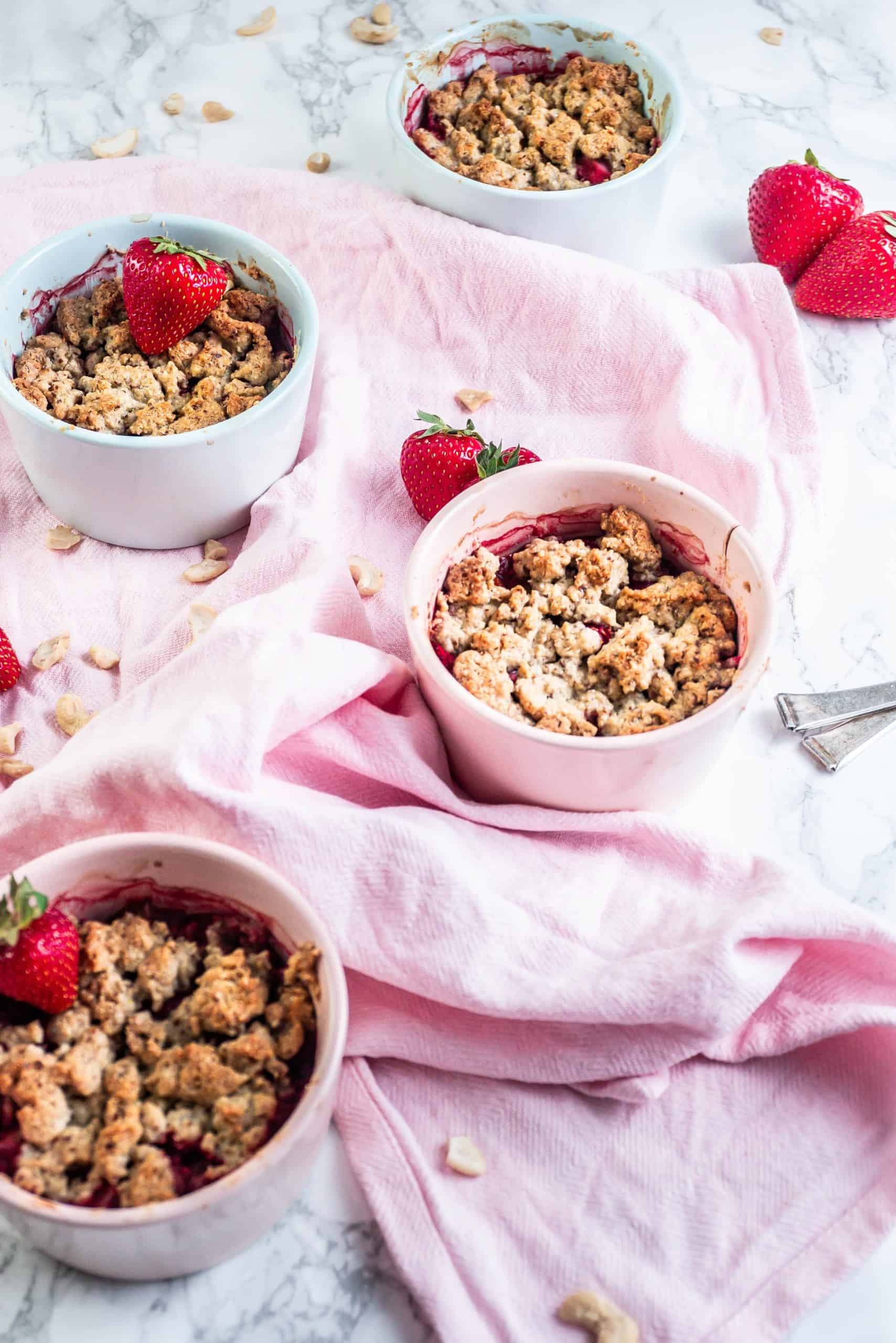 Strawberry Rhubarb Crumble in small ramekins with strawberries and cashews strewn around with four visible but one being in focus