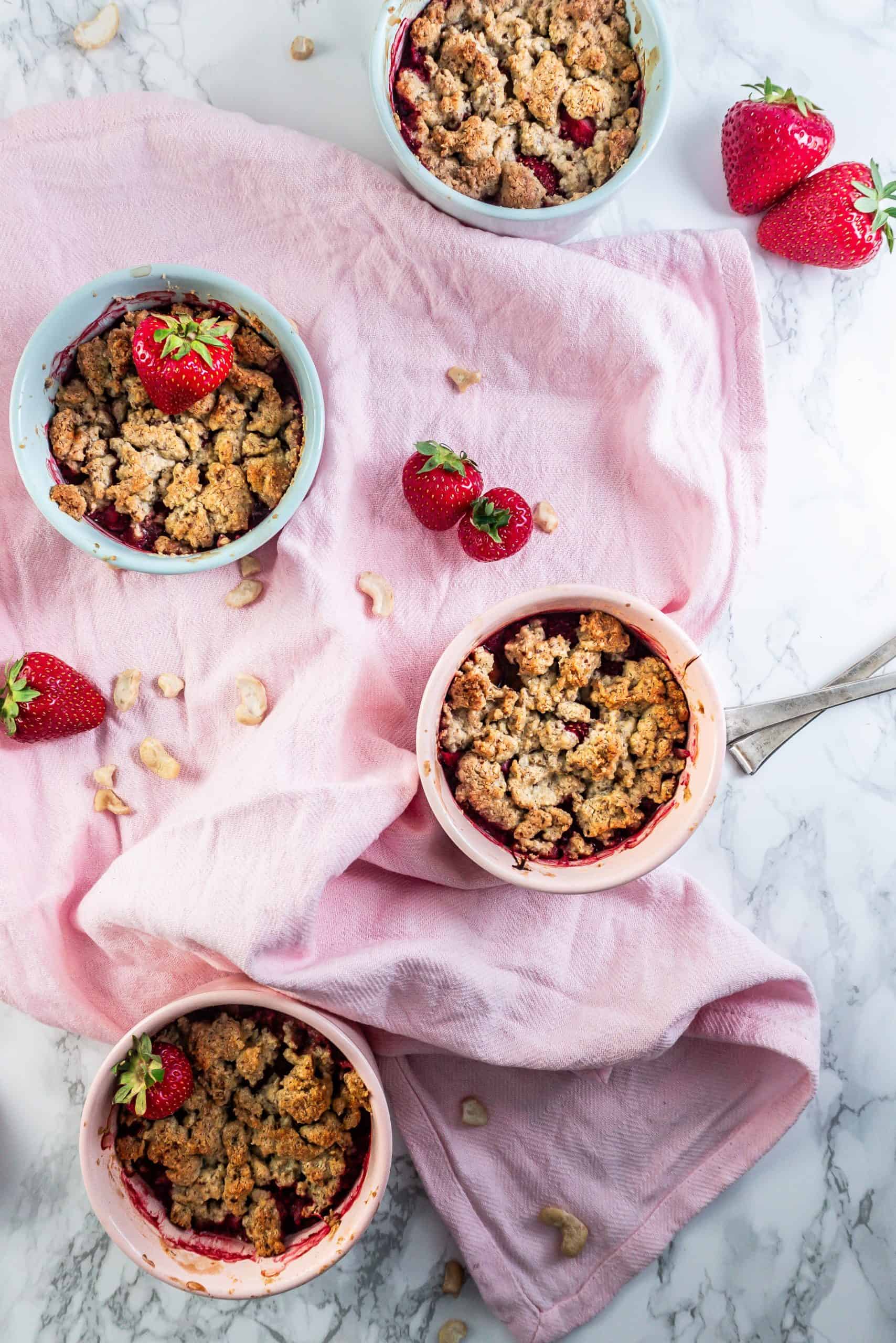 Strawberry Rhubarb Crumble in small ramekins with strawberries and cashews strewn around; photographed from above