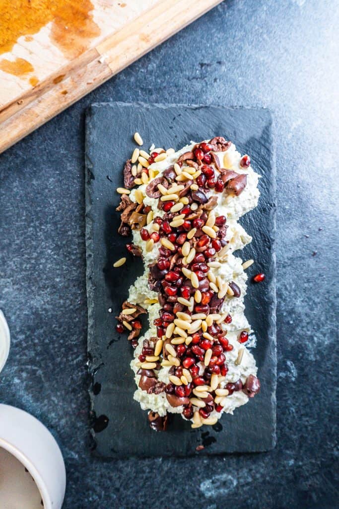 Preparing the creamy whipped feta dip on a serving platter with olives, pomegranate seeds, and pine nuts already sprinkled on top