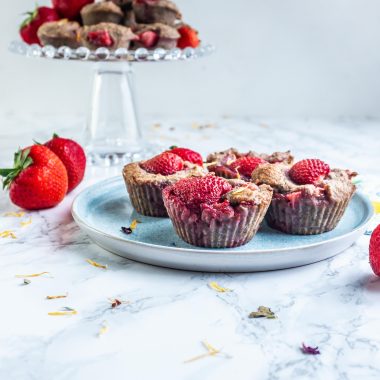 Strawberry Banana Muffins on a plate with some smaller muffins visible on a cake stand in the back, surrounded by strawberries