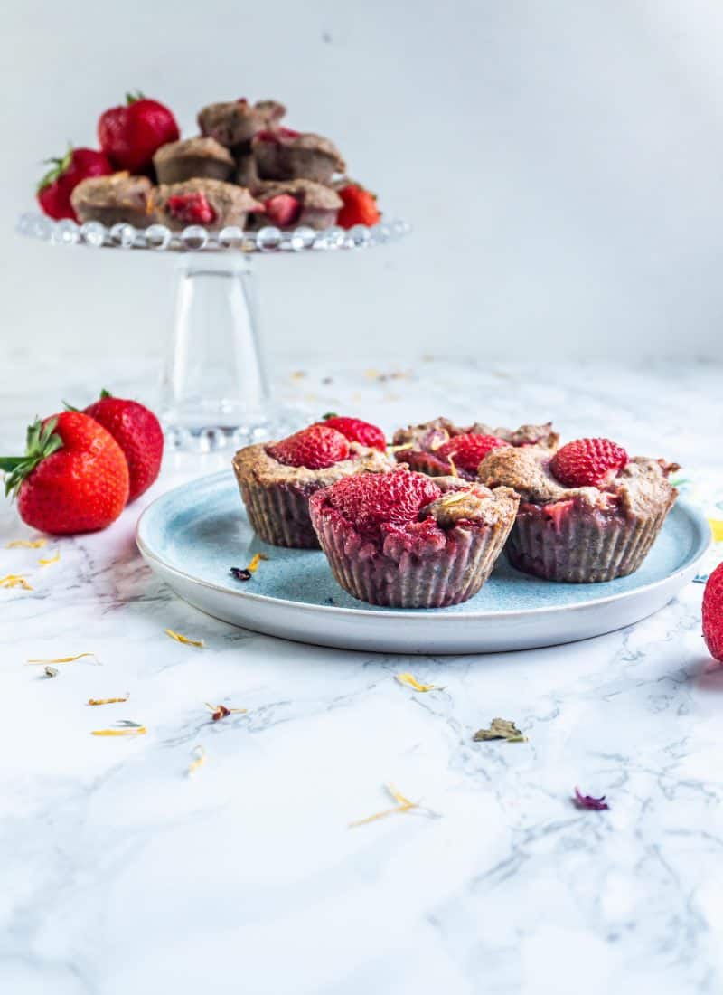 Strawberry Banana Muffins on a plate with some smaller muffins visible on a cake stand in the back, surrounded by strawberries