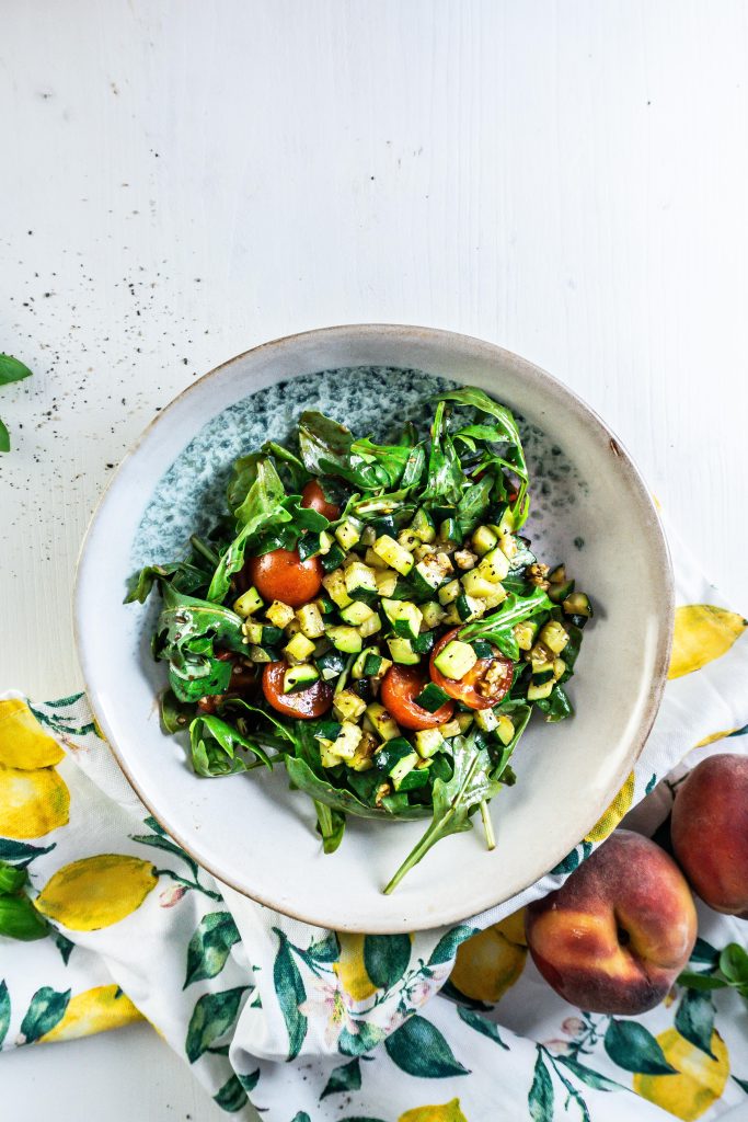 Cherry tomatoes, rocket, basil, and walnuts in a bowl with a balsamic glaze added in a bowl, with grilled courgette added, and ready to add additional ingredients for the Grilled Peach Salad with Burrata