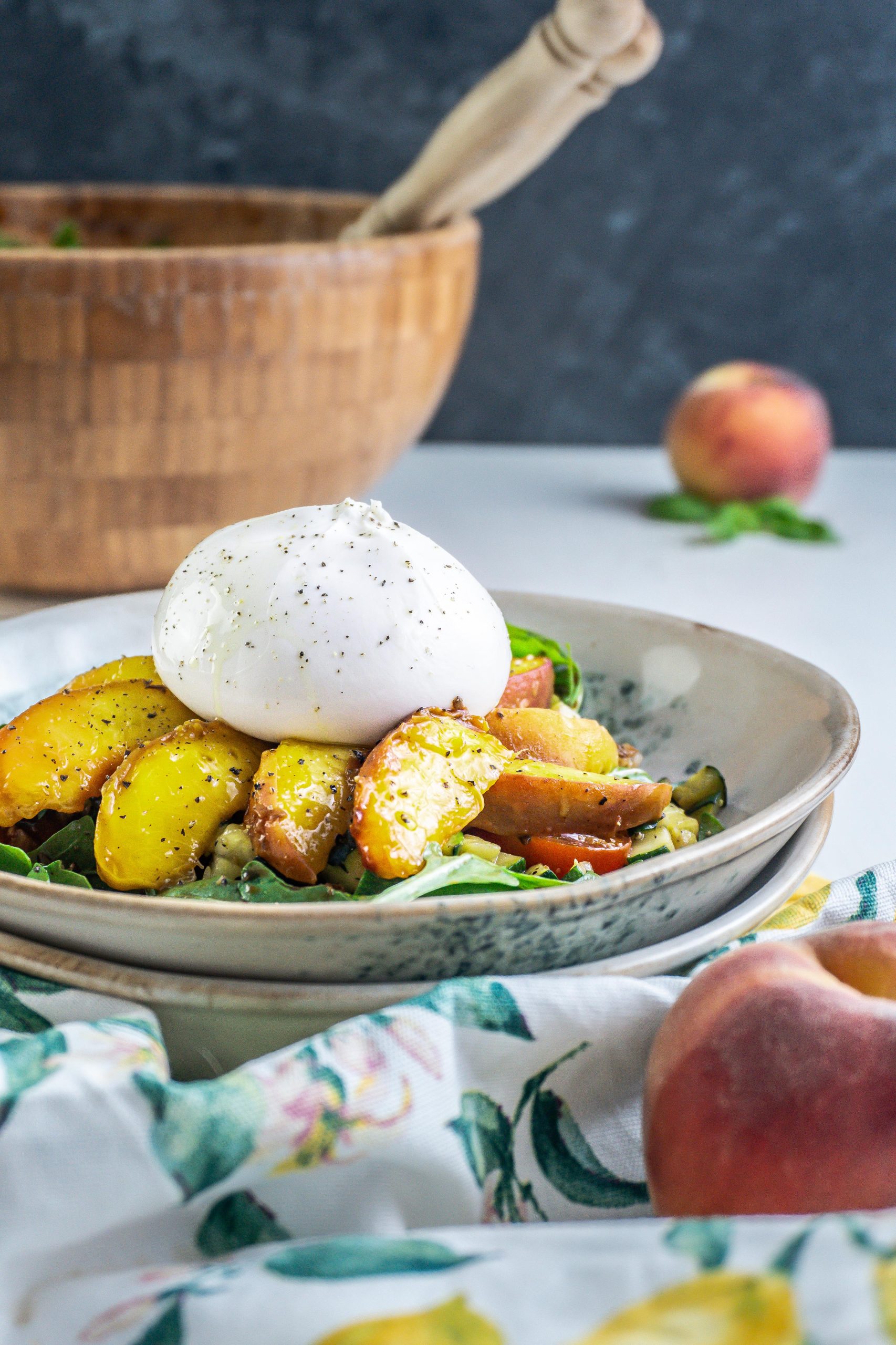 Grilled Peach Salad with Burrata on a plate with a kitchen towel, peaches, and basil visible in the background; photographed from a side-angle