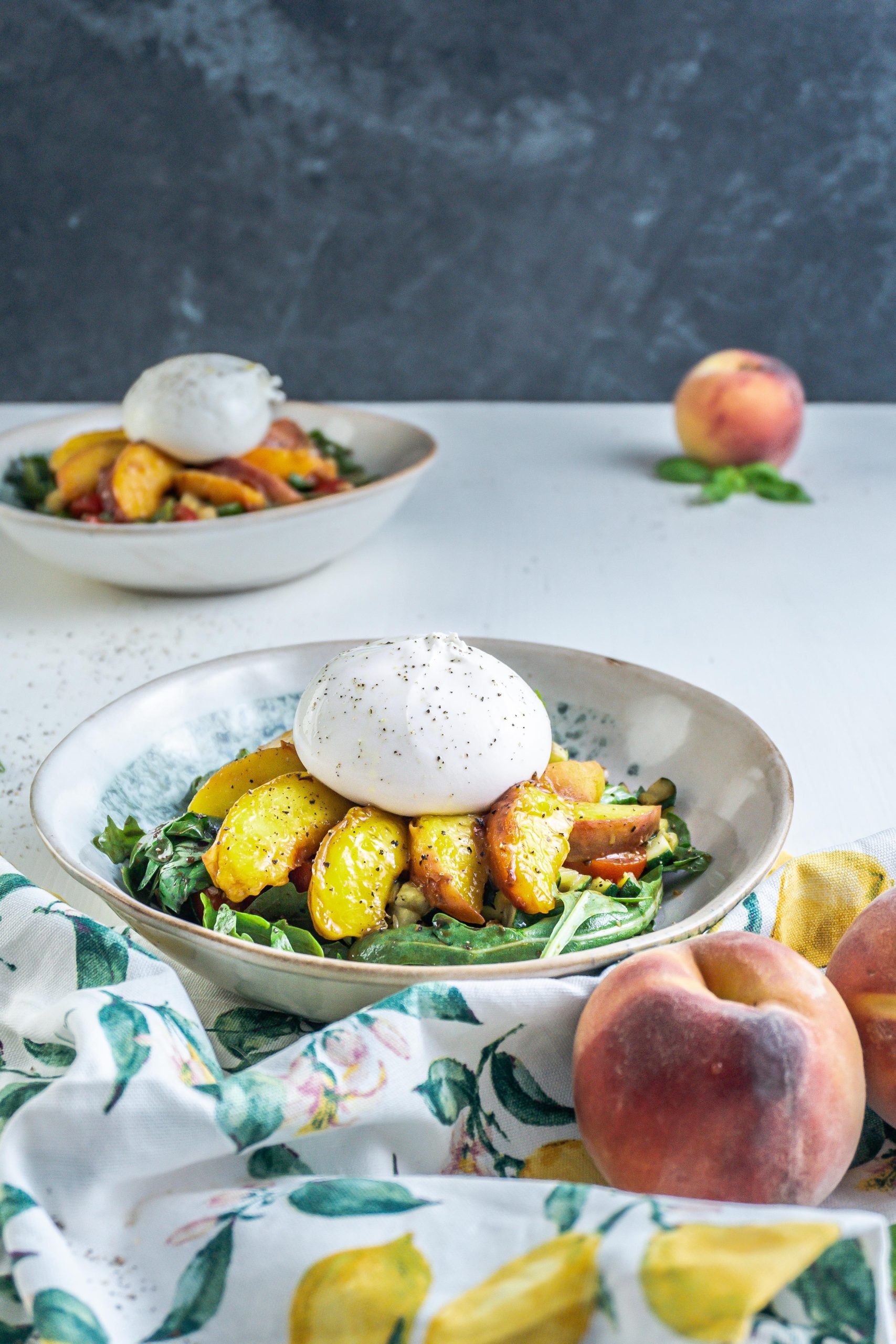 Grilled Peach Salad with Burrata on a plate with a kitchen towel, peaches, and basil visible in the background and a second plate in the background