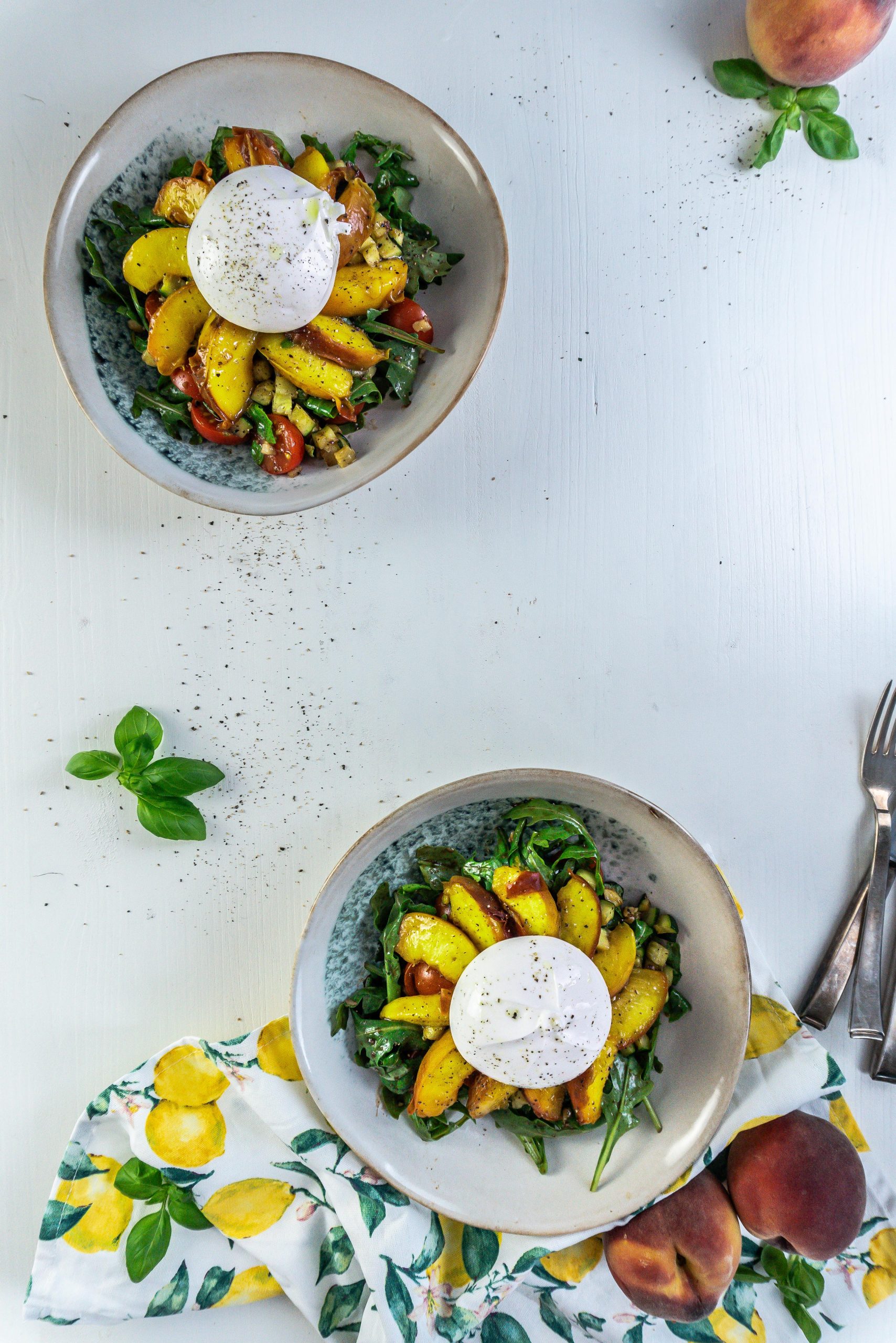 Grilled Peach Salad with Burrata on a plate with a kitchen towel, peaches, and basil visible in the background, and a second plate in the background; photographed from above