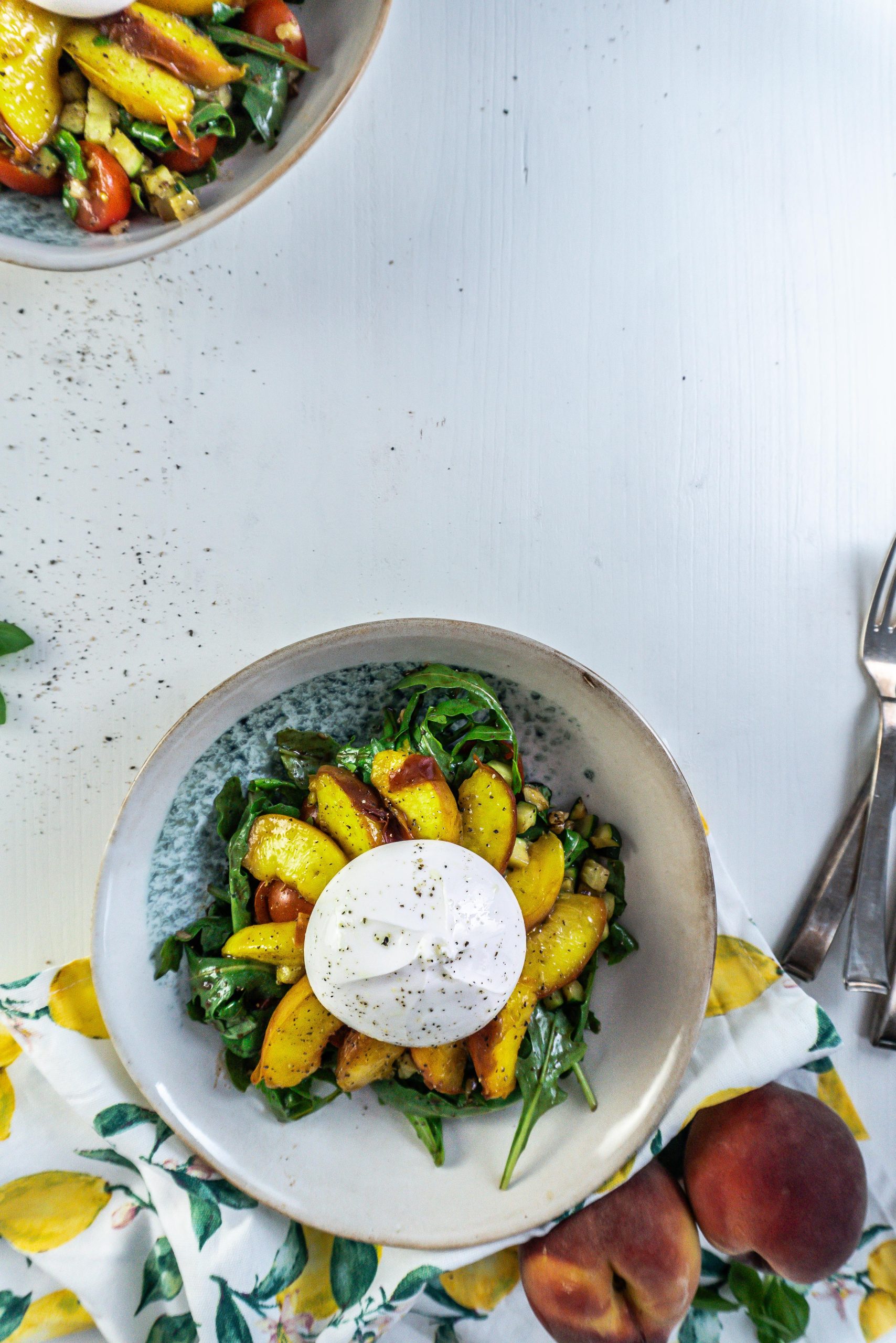 Grilled Peach Salad with Burrata on a plate with a kitchen towel, peaches, and basil visible in the background and a second plate in the background