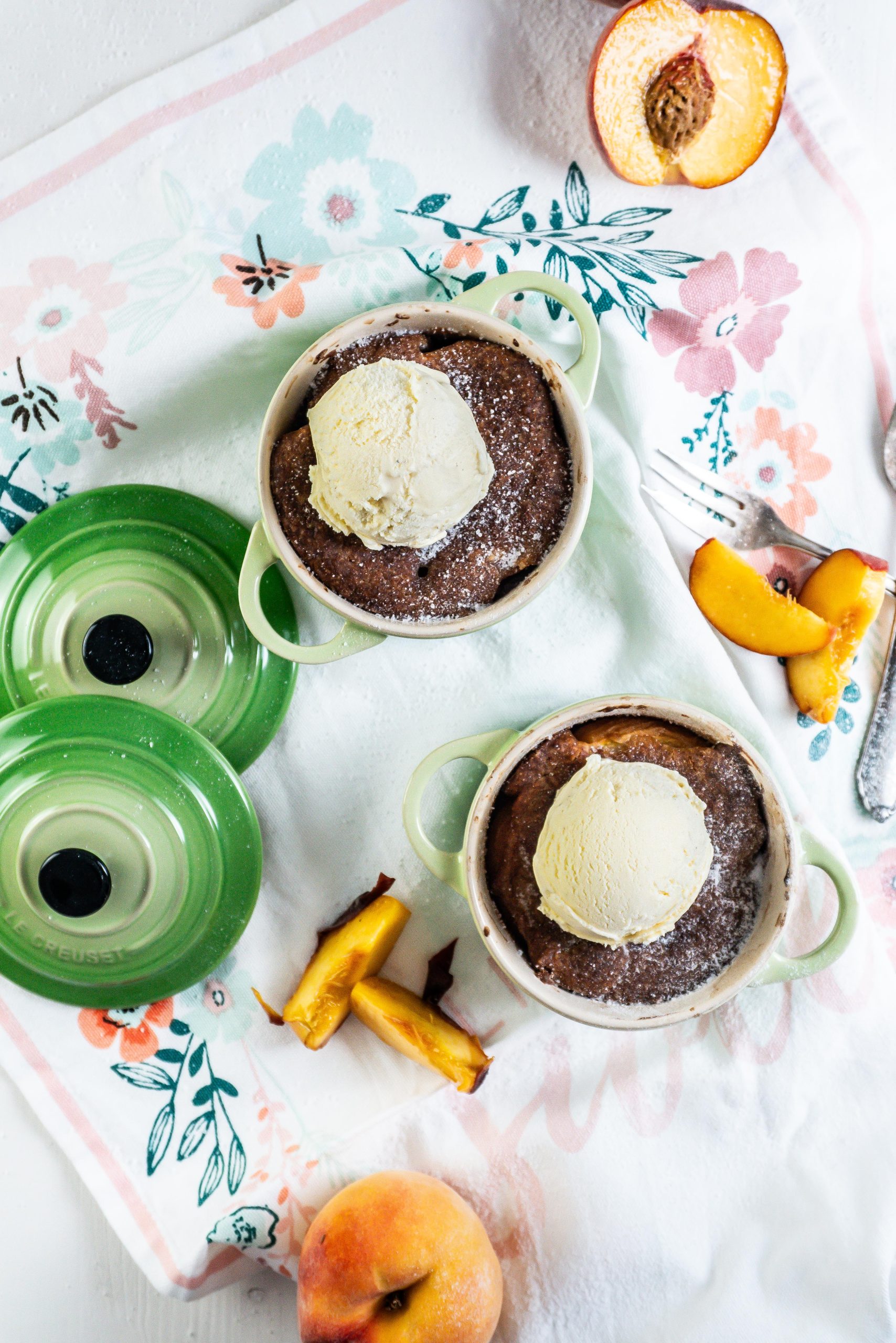 Mini Peach Cobblers with ice cream on top in small ramekins with peaches strewn around and two forks lying on the side